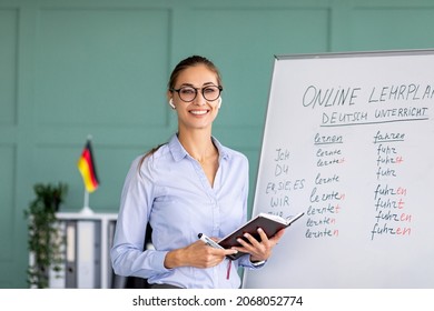 Portrait of cheerful German teacher standing near blackboard, conducting internet lesson and smiling at camera. Positive tutor giving language class on video call or web conference