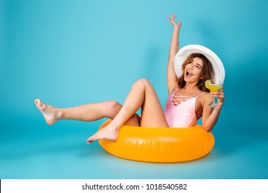Portrait of a cheerful girl dressed in swimsuit sitting on inflatable ring and holding cocktail isolated over blue background