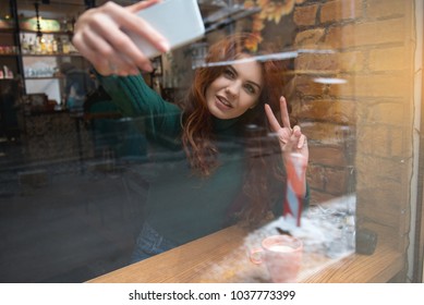 Portrait of cheerful girl photographing herself on mobile phone and smiling. She is showing peace sign not camera while resting in cozy cafeteria. View from glass window - Powered by Shutterstock