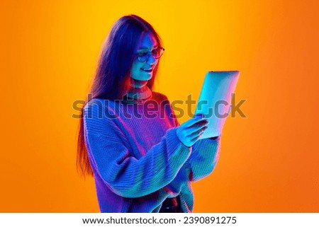 Portrait of cheerful girl, student dressed wool sweater holding tablet and chatting online in neon light against gradient background. Concept of beauty, human emotions, shopping, sale, fashion, style.