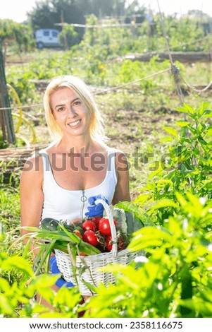 Portrait of cheerful girl in tank top and jean cutoffs posing with fresh vegetable harvest in backyard garden.