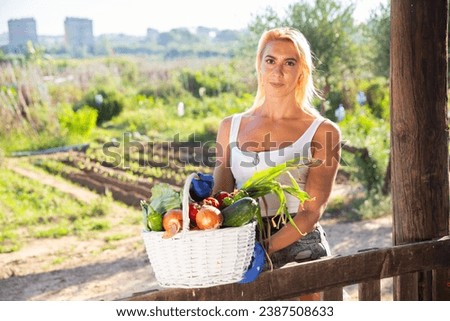 Portrait of cheerful girl in tank top and jean cutoffs posing with fresh vegetable harvest in backyard garden.