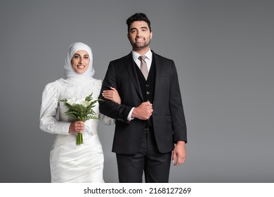 portrait of cheerful groom in suit standing muslim bride with wedding bouquet of calla lily flowers isolated on grey