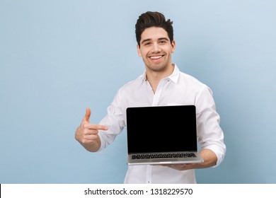 Portrait Of A Cheerful Handsome Young Man Standing Isolated Over Blue Background, Working On Laptop Computer, Showing Blank Screen Laptop