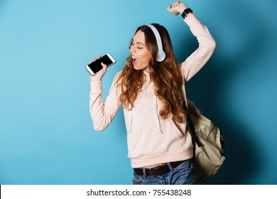 Portrait of a cheerful happy girl student with backpack listening to music with headphones while showing blank screen mobile phone and dancing isolated over blue background