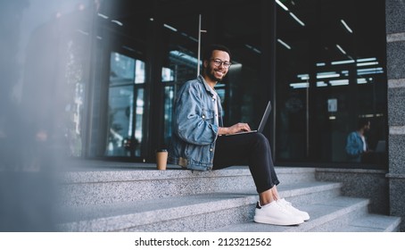 Portrait of cheerful hipster guy in optical glasses for provide eyes correction smiling at camera during time for doing distance job on netbook, happy male blogger with laptop posing at urban stairs - Powered by Shutterstock