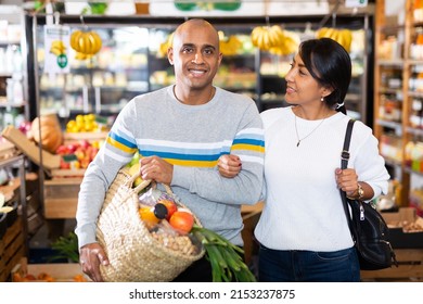 Portrait Of Cheerful Hispanic Couple Shopping In Grocery Store Carrying Wicker Basket Full Of Fresh Food Goods