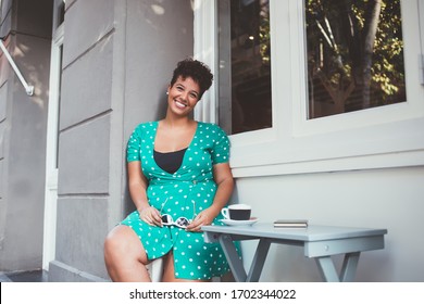 Portrait of cheerful Hispanic woman dressed in stylish clothing apparel resting in sidewalk cafe and smiling at camera during free time outdoors, happy hipster girl in dress in polka dots rejoicing