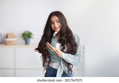 Portrait Of Cheerful Indian Lady Brushing Her Beautiful Long Hair, Using Wooden Brush At Home. Positive Young Woman Taking Care Of Her Wavy Locks. Domestic Spa Salon, Beauty Concept