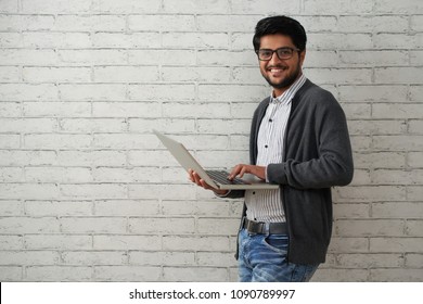 Portrait Of Cheerful Indian Programmer With Laptop Standing Against White Brick Wall