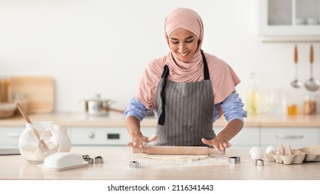 Portrait Of Cheerful Islamic Housewife Wearing Hijab And Apron Baking In Kitchen, Happy Young Muslim Woman Rolling Out Dough On Table, Preparing Cookies At Home, Panorama With Copy Space - Powered by Shutterstock