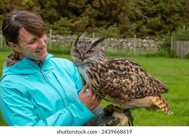 PORTRAIT: Cheerful lady with a beautiful Eurasian eagle owl perched on her arm. Female traveller is having an authentic experience while on an exciting tourist visit to a Scottish falconry center. - Powered by Shutterstock