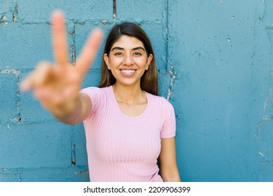 Portrait Of A Cheerful Latin Woman Smiling Looking At The Camera Making The Peace Sign Against A Blue Wall Outdoors