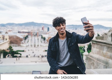 Portrait Of Cheerful Latino Male Traveller Resting During Spanish Vacations To Barcelona Clicking Selfie Pictures On Montjuic Area, Millennial Man Photographing Himself With Cityscape On Background