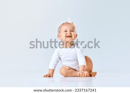 Portrait of cheerful little baby girl, toddler, child calmly sitting and laughing against grey studio background. Concept of childhood, family, newborn lifestyle, happiness, care. Copy space for ad