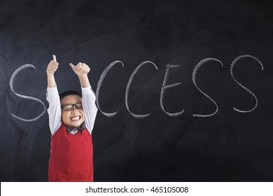 Portrait of a cheerful little boy showing thumbs up with Success word on the blackboard, shot in the class
