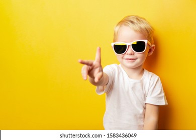 Portrait of cheerful little boy in a white t-shirt wearing sunglasses and looking away on orange background showing two fingers peace sign.