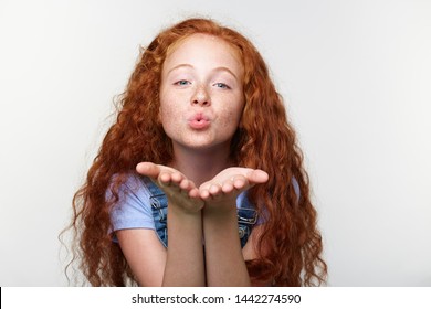 Portrait Of Cheerful Little Girl With Ginger Hair And Freckles, Send Kiss At The Cam, Looks Happy, Standing Over White Wall.