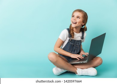 Portrait of a cheerful little girl isolated over blue background, studying with laptop computer