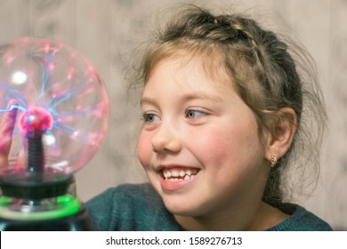 Portrait of a cheerful little girl playing with a plasma ball. The child smiles and laughs while looking at the lightning in a decorative round lamp. Selective focus.