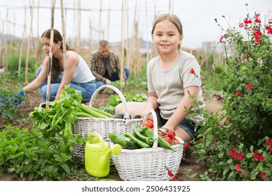 Portrait of cheerful little girl posing with freshly harvested greens and vegetables in homestead, her parents gardening in background - Powered by Shutterstock