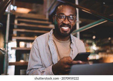 Portrait of cheerful male blogger with cellphone gadget and cropped laptop smiling at camera during free time, happy African American man in classic glasses using smartphone and netbook indoors - Powered by Shutterstock
