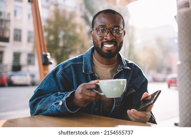 Portrait Of Cheerful Male Blogger In Optical Eyewear Holding Modern Cellphone Device And Coffee Cup, Happy Millennial Hipster Guy With Digital Mobile Technology Smiling At Camera In Sidewalk Cafe