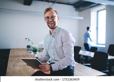 Portrait of cheerful male entrepreneur holding digital tablet and laughing at camera during work day in company office, satisfied businessman in optical eyeglasses rejoicing at table desktop