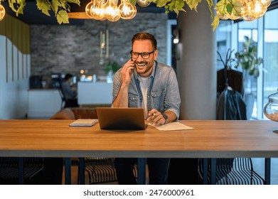 Portrait of cheerful male entrepreneur making phone call and working over laptop on desk in office - Powered by Shutterstock