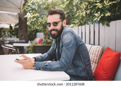 Portrait of cheerful male generation Z spending leisure day in comfortable street cafeteria with modern cellphone for blogging and networking, happy successful man smiling at camera during free time