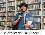 Portrait of cheerful male international Indian student with backpack, learning accessories standing near bookshelves at university library or book store during break between lessons. Education concept