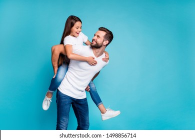 Portrait Of Cheerful Man Holding His Kid With Brunette Hairstyle Piggyback Wearing White T-shirt Denim Jeans Isolated Over Blue Background