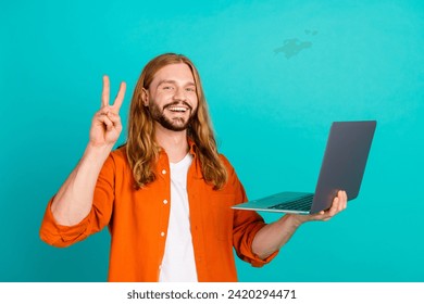 Portrait of cheerful man with long hairstyle dressed orange shirt hold laptop showing v-sign symbol isolated on teal color background