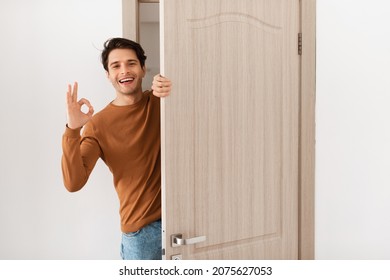 Portrait of cheerful man standing in doorway of modern apartment, millennial male homeowner holding door looking out and smiling, greeting visitor, showing okay sign gesture, approving - Powered by Shutterstock