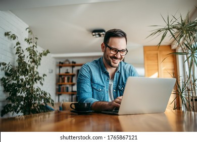 Portrait Of A Cheerful Man Using Laptop While Sitting At Wooden Table At Cozy Home.