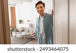 Portrait of cheerful man welcoming inviting visitor to enter his home, happy young guy standing in doorway of modern apartment, millennial male holding door looking out showing living room with hand