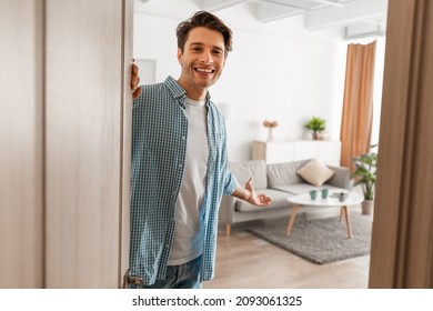 Portrait of cheerful man welcoming inviting visitor to enter his home, happy young guy standing in doorway of modern apartment, millennial male holding door looking out showing living room with hand - Shutterstock ID 2093061325