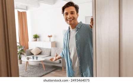 Portrait of cheerful man welcoming inviting visitor to enter his home, happy young guy standing in doorway of modern apartment, millennial male holding door looking out showing living room with hand - Powered by Shutterstock