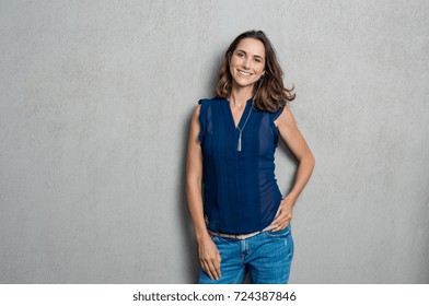 Portrait of cheerful mature woman standing against grey wall. Happy mid woman looking at camera against grey background with copy space. Smiling carefree latin woman looking at camera.