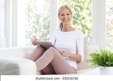 Portrait Of Cheerful Middle Age Woman Sitting At Sofa And Using Her Digital Tablet While Working Online At Home. 
