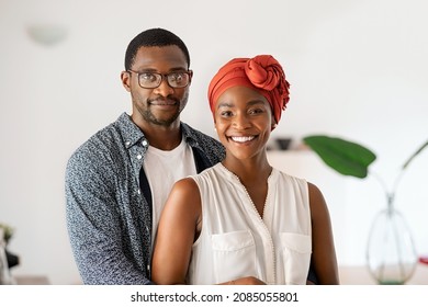 Portrait of cheerful middle aged couple embracing at home. Romantic mid adult black man hugging beautiful wife with traditional red turban while smiling and looking at camera. Mature african couple.
