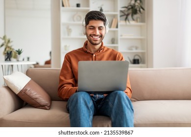 Portrait Of Cheerful Middle Eastern Guy Working With Laptop At Home, Smiling Arab Millennial Man Sitting On Couch In Living Room And Using Pc Computer, Listening To Music In Wireless Headset