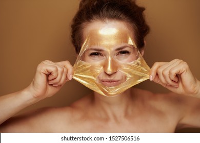 Portrait Of Cheerful Modern 40 Year Old Woman Removing Golden Cosmetic Face Mask Isolated On Brown Background.