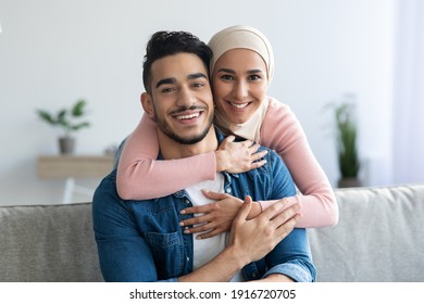 Portrait of cheerful muslim couple embracing at home, copy space, closeup. Smiling young woman in headscarf hugging her loving husband from behind, middle-eastern family spending time together