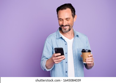 Portrait of cheerful positive man have free time relax after work use smartphone check social network hold mug with hot beverage wear denim jeans shirt isolated over violet color background - Powered by Shutterstock