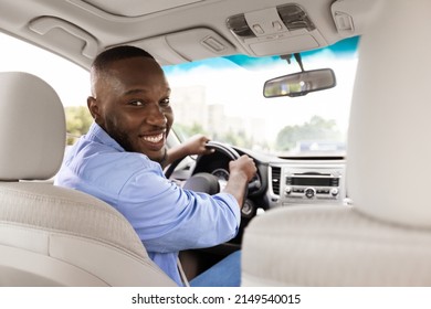 Portrait Of Cheerful Positive Young Black Male Driver Turning Looking Back At Camera Sitting In A New Luxury Car, View From Rear Seat Passenger. Excited Guy Riding Holding Hands On Steering Wheel