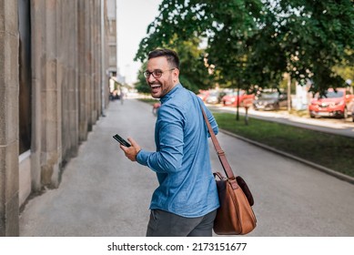 Portrait of cheerful professional commuter looking over shoulder. Young male executive is carrying laptop bag. He is walking on sidewalk by building in the city. - Powered by Shutterstock