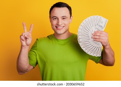 Portrait of cheerful rich guy show v-sign hold money fan on yellow background