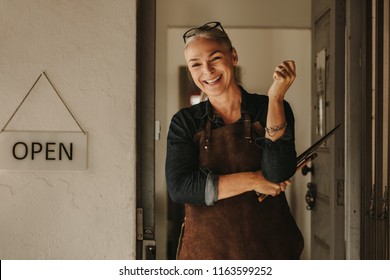 Portrait Of Cheerful Senior Female Jeweler Standing At Workshop Door. Woman Jewelry Maker In Apron Holding Tools At Workshop Entrance.