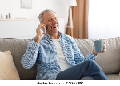 Portrait Of Cheerful Senior Man Talking On Cell Phone Sitting On Couch At Home In Living Room, Making Informal Call With Friends Or Family, Discussing Something Drinking Coffee. Remote Communication